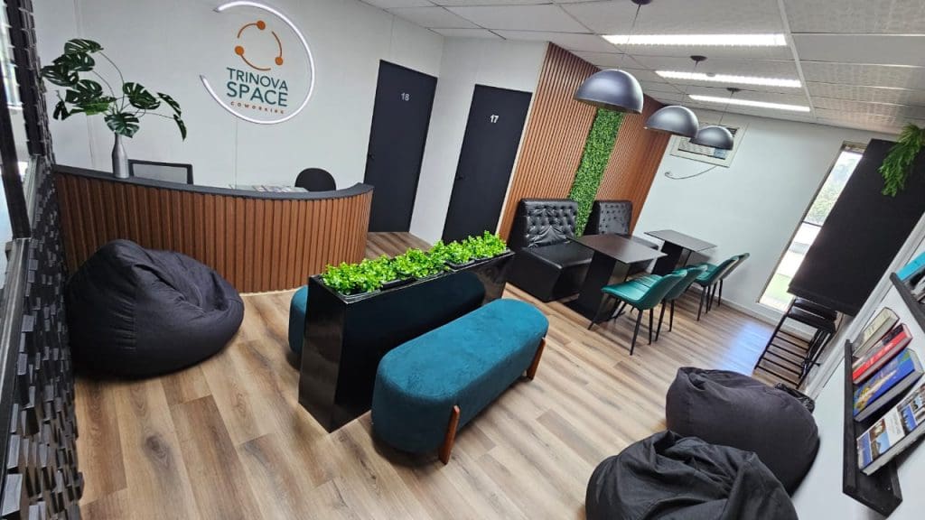 Lounge com bancos azul-petróleo, jardineira, pufes e balcão de madeira com o logo Trinova Space